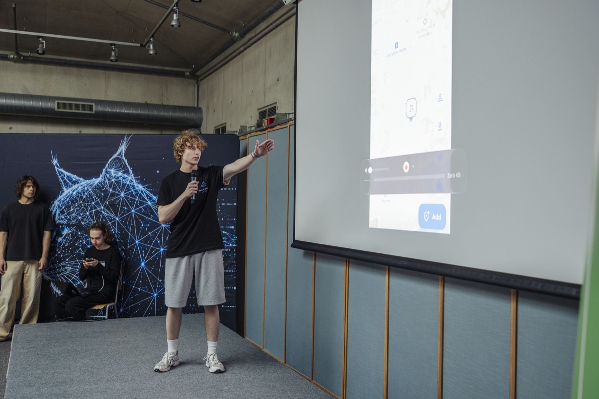 Viktor Vokh on stage with a microphone, presenting the deVoda app on the projector screen behind him at the Day One Ukraine Hackathon