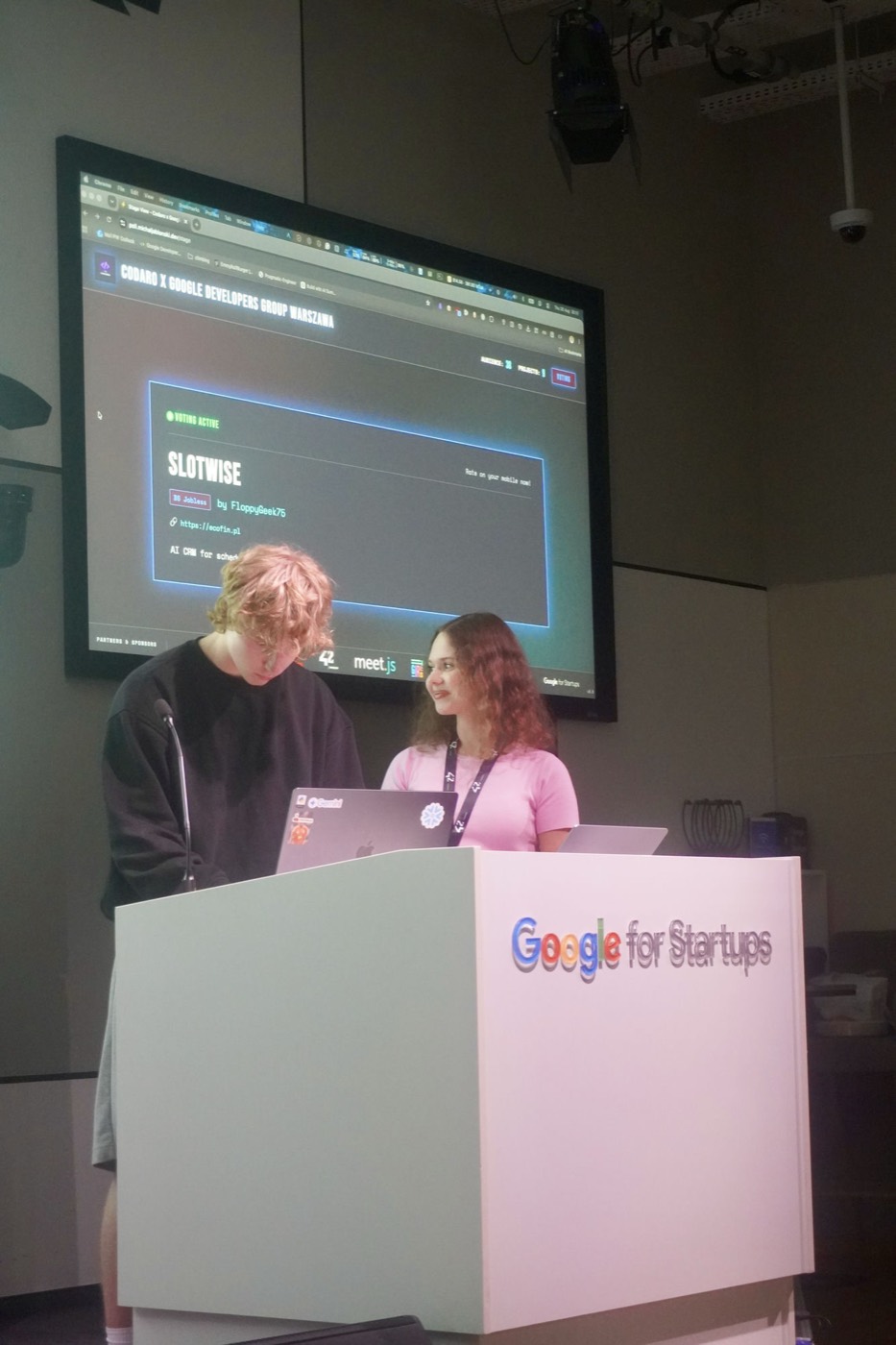 Two of the team at the Google for Startups podium as Slotwise is put up for audience voting on the screen behind them